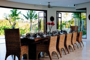 A dining area with a long table, wicker chairs, and a view of greenery outside. - Umah Daun Villa by Nakula (Bali)