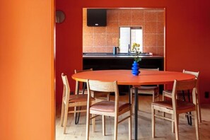 A dining area with a round wooden table and six chairs, a blue vase with flowers, and a kitchen in the background with orange tiles and a black TV mounted on the wall. - VILLA + SECCA (Ito)