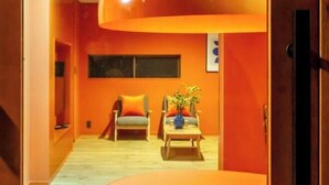 A room with orange walls, a wooden floor, a sofa with orange cushions, a coffee table with a vase, and a window. - VILLA + SECCA (Ito)