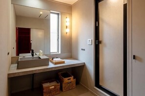 A bathroom with a large mirror, a sink, and a window. There are wooden baskets under the sink and a door to the right. - VILLA + SECCA (Ito)