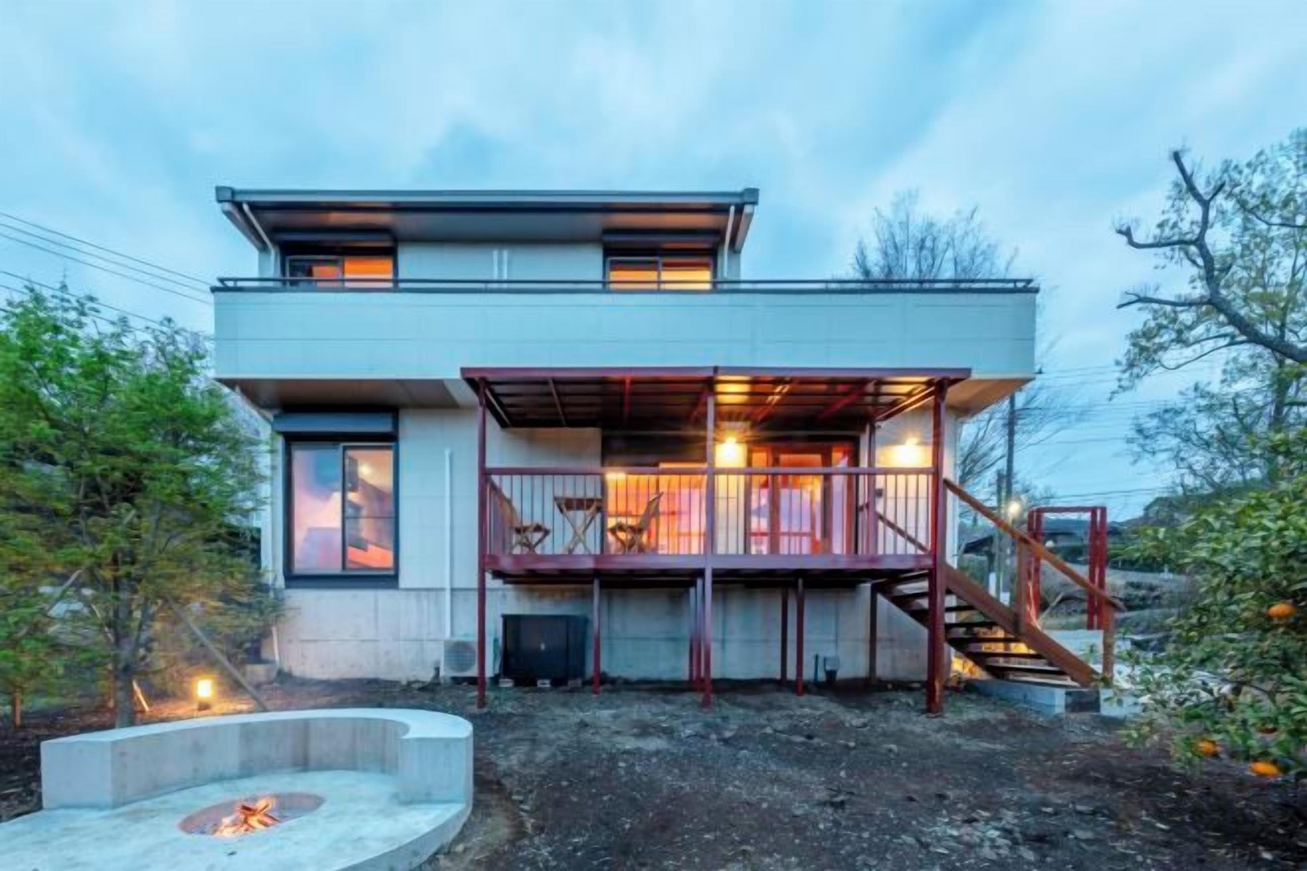Egy modern, kétszintes ház, fedett kültéri terasszal, tűzrakóhellyel és egy lépcsővel, amely a felső szintre vezet.