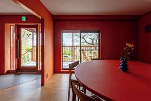 A room with a red table, wooden chairs, a vase with flowers, and a large window with a view of the outdoors. - VILLA + SECCA (Ito)