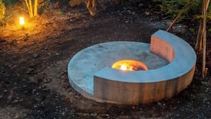 A circular concrete fire pit with a central fire, surrounded by a natural outdoor setting with trees and a small light source to the left. - VILLA + SECCA (Ito)