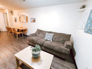 A living room with a brown sofa, a wooden coffee table with a plant, a dining table with chairs, and a white wall with framed pictures.