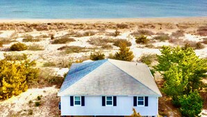A house with a gabled roof, located on a sandy beach with sparse vegetation and a view of the ocean.