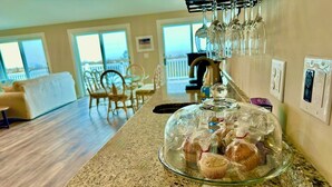 A kitchen counter with a glass dome displaying muffins, a dining area with chairs and a table, and a view of a balcony through large windows.