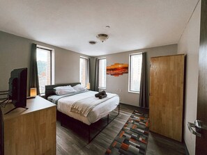 A hotel room with a bed, a desk, a television, and a window. - 2BR 2BA Downtown Apt | Water + City Views (Pittsburgh)