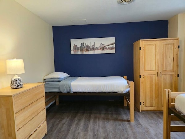 A single bed with a headboard, a wooden wardrobe, a bedside table with a lamp, and a cityscape painting on the wall.