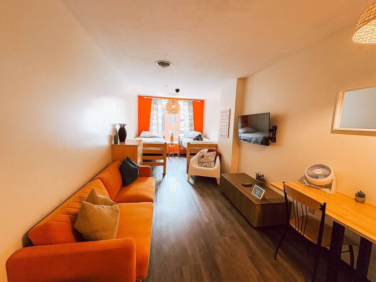 A compact living space with a sofa, a small dining table, a TV mounted on the wall, and a window with curtains.
