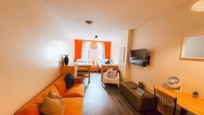 A compact living space with a sofa, a small dining table, a TV mounted on the wall, and a window with curtains.