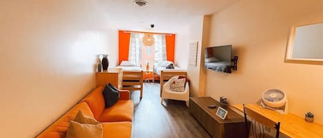 A compact living space with a sofa, a small dining table, a TV mounted on the wall, and a window with curtains.