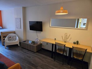 Une chambre d’hôtel moderne équipée d’un téléviseur à écran plat, d’une table à manger en bois avec des chaises, d’une petite table d’appoint avec un ventilateur, d’un tapis rouge et d’un grand miroir mural.