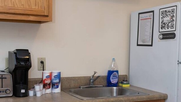 A kitchen with wooden cabinets, a stainless steel sink, a coffee maker, and a refrigerator with a QR code. - Large Private Downtown Apartment | 2BR + 2BA + 60” (Pittsburgh)