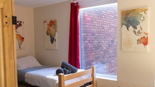 A single bed with a wooden headboard, a red curtain, and two wall art pieces depicting maps. - Large Private Downtown Apartment | 2BR + 2BA + 60” (Pittsburgh)
