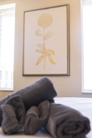 A framed botanical print on a wall, a rolled gray towel on a bed, and a window with blinds.