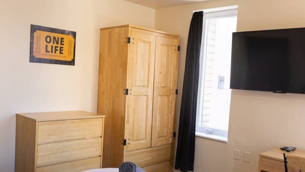 A bedroom with a bed, a wooden wardrobe, a desk, a television, and a window with curtains.