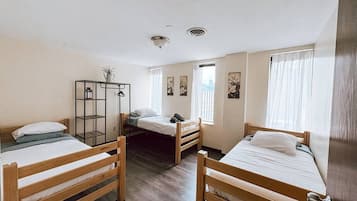 A dormitory room with three wooden bunk beds, a metal shelf unit, and a window with sheer curtains.
