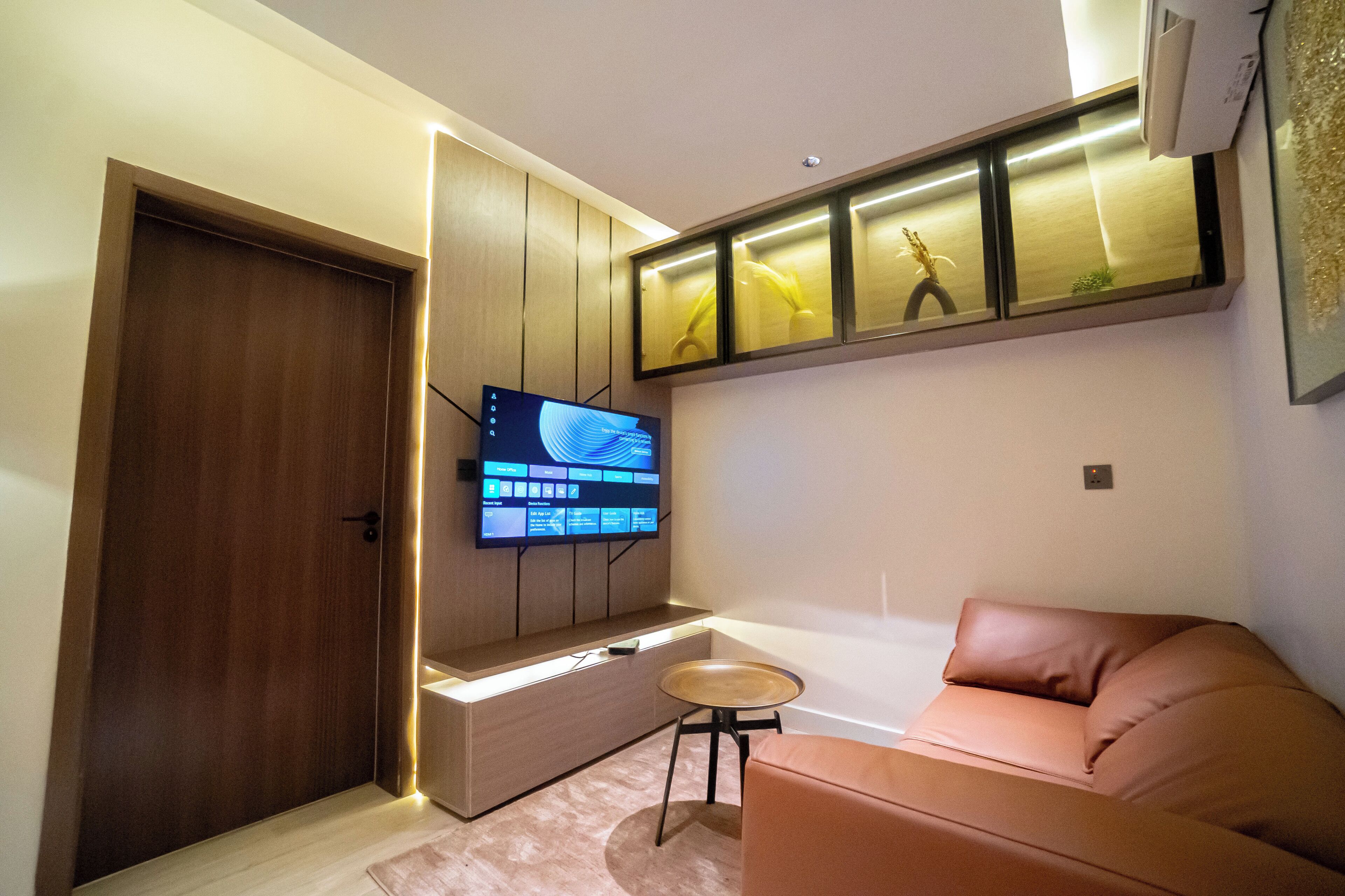 Un salon moderne avec un téléviseur à écran plat fixé au mur, un canapé en cuir marron, une petite table ronde et une porte en bois.