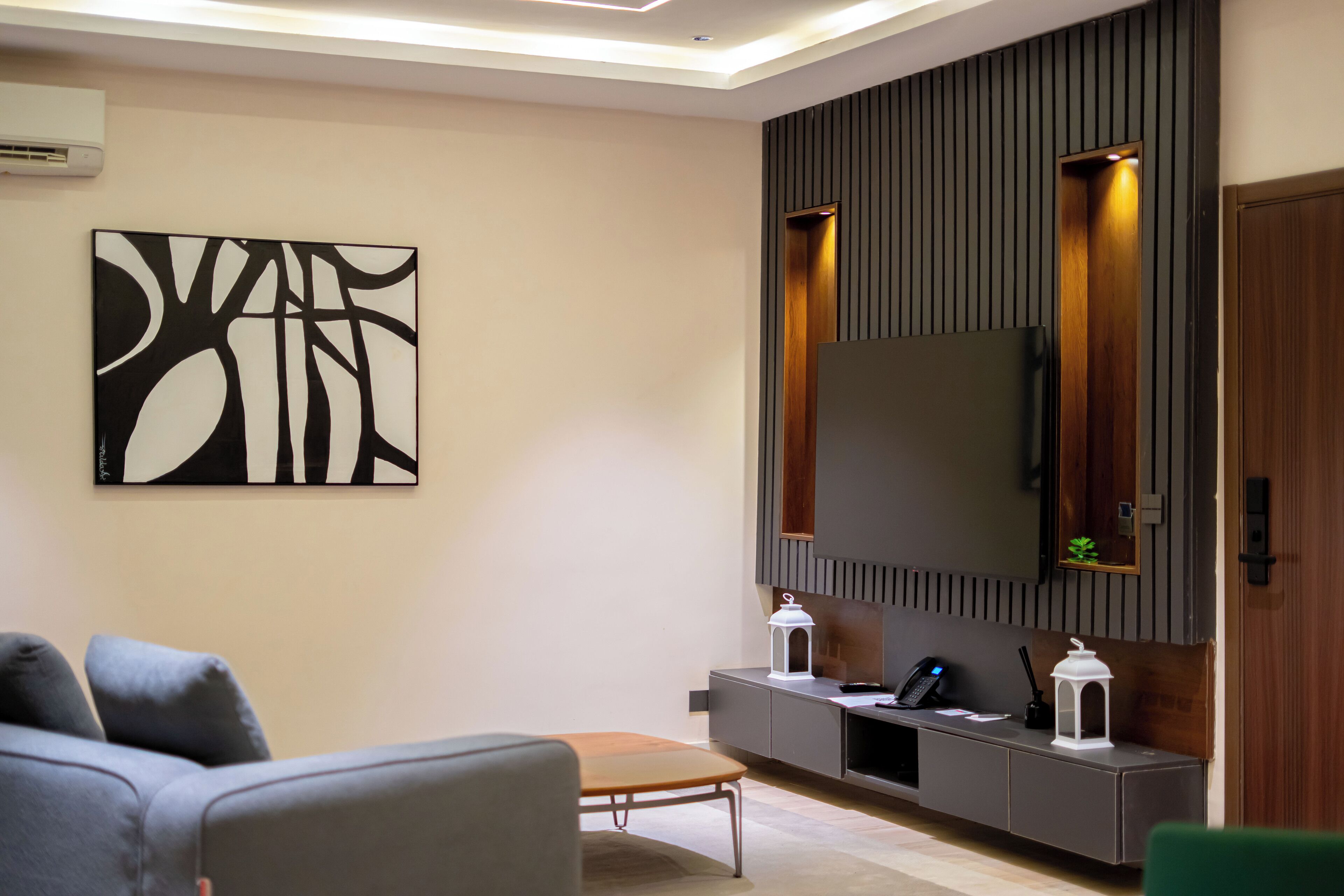A modern living room with a flat-screen TV, a large abstract painting, a sofa, a coffee table, and a console table with a phone and a lantern.