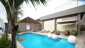 A swimming pool with lounge chairs and umbrellas, surrounded by a modern building with a covered patio area. - GREYFIELD HOTEL AND APARTMENTS (Benin City)