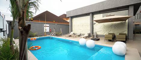 A swimming pool with lounge chairs and umbrellas, surrounded by a modern building with a covered patio area.