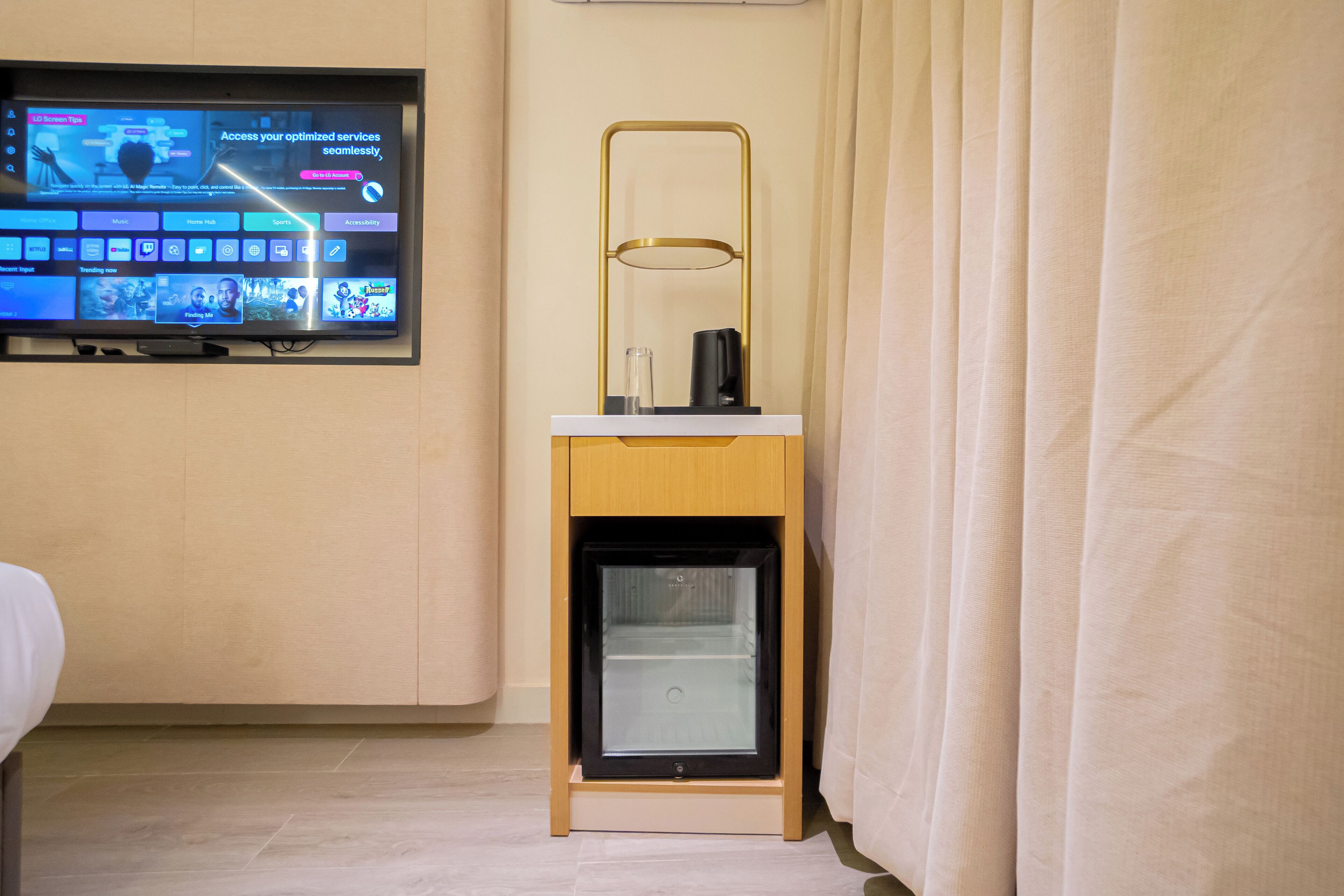 A hotel room with a television mounted on the wall displaying various channels and a small refrigerator on a wooden stand.