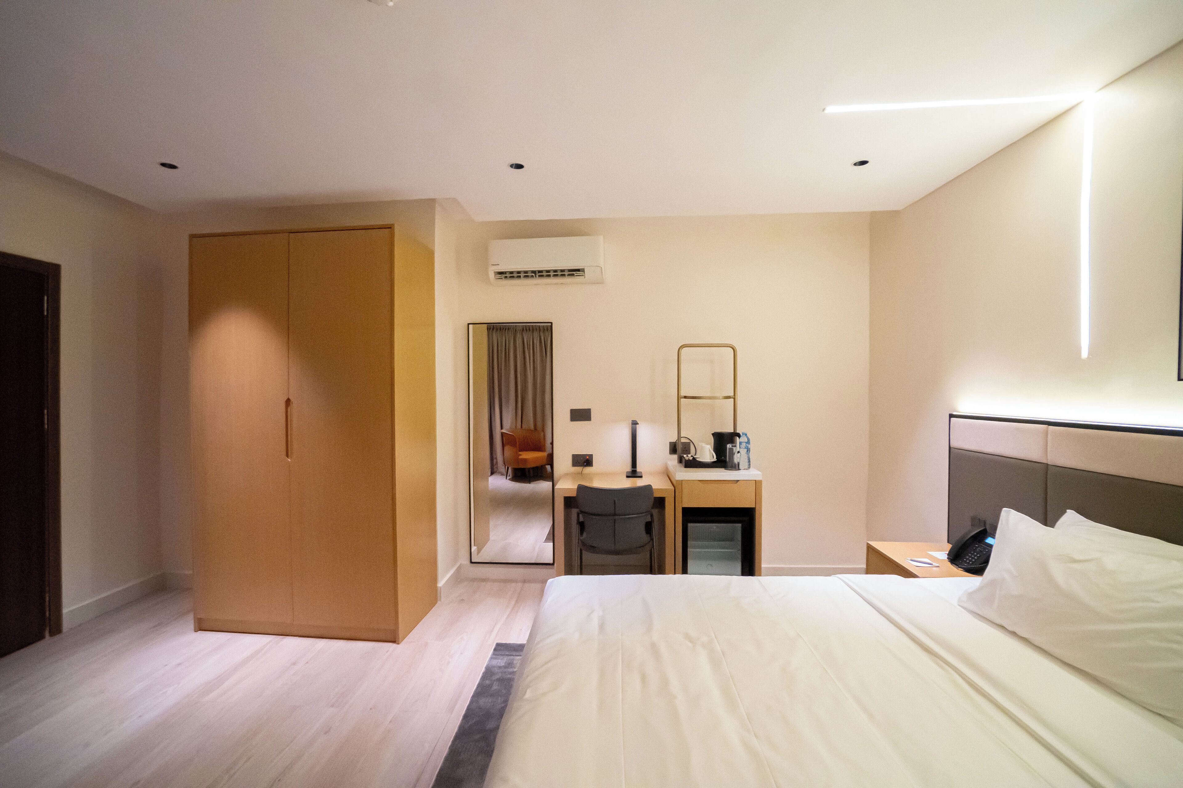 A modern hotel room with a large bed, a desk with a chair, a wardrobe, a mirror, and an air conditioning unit.