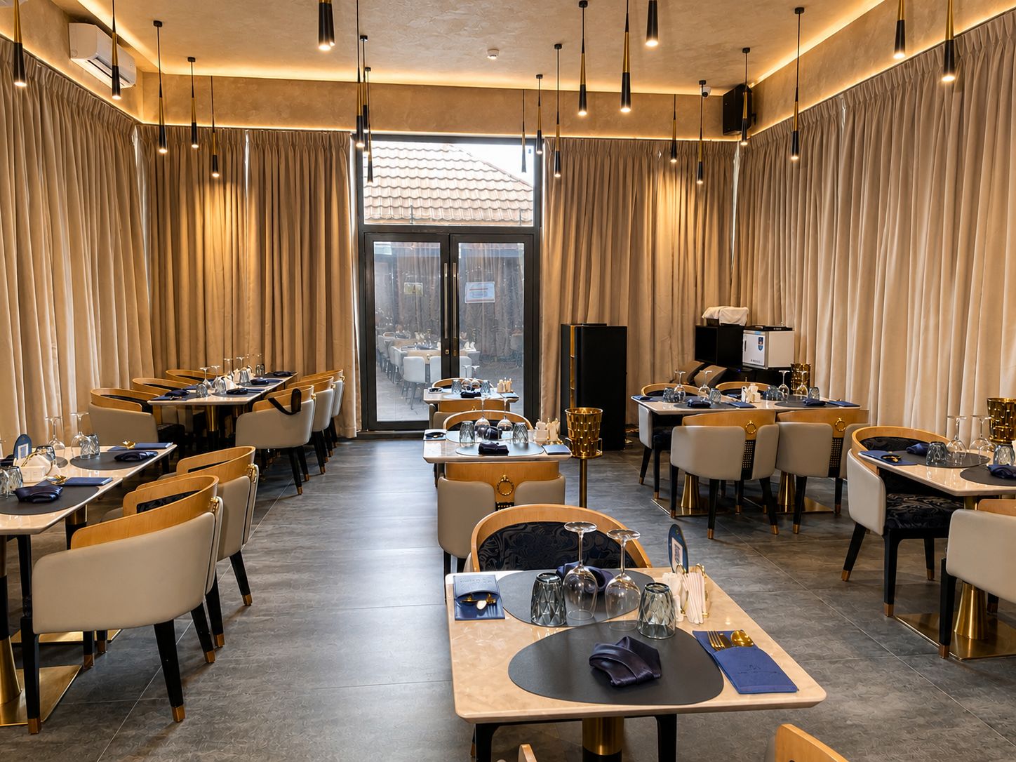 Un restaurante con varias mesas y sillas, un amplio ventanal con cortinas y lámparas colgantes.