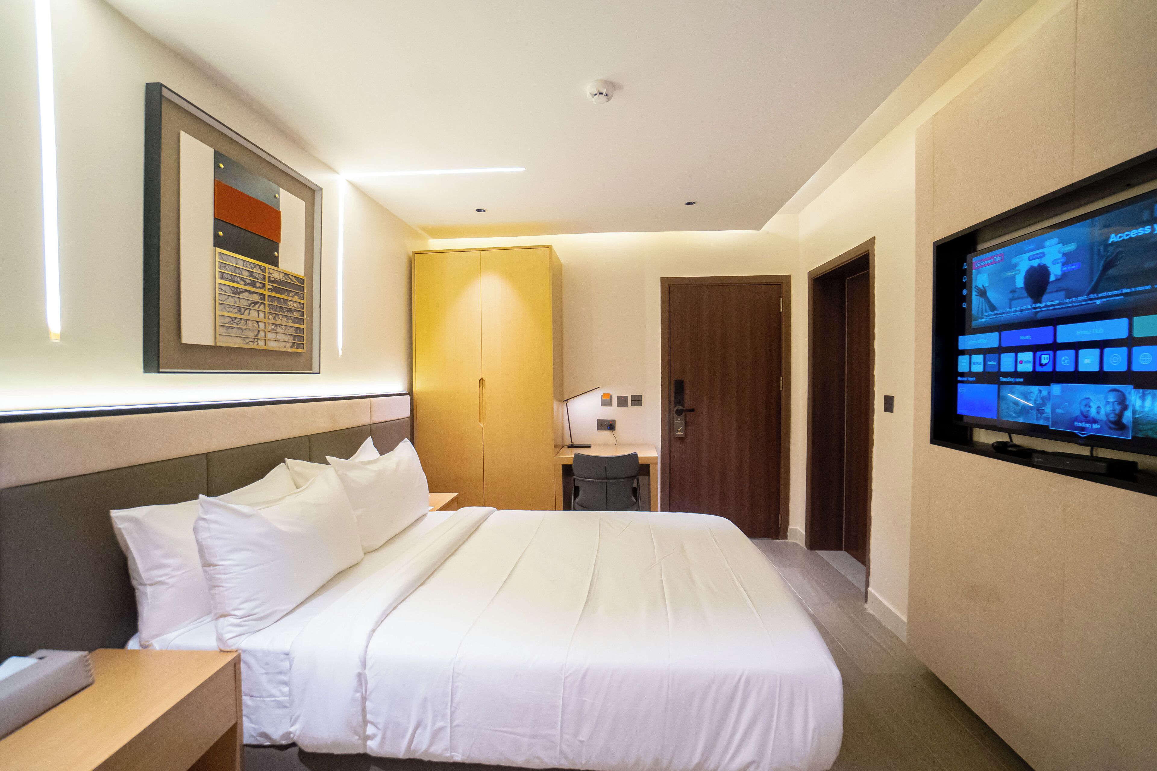 A hotel room with a large bed, a desk, a television, and a framed artwork on the wall.