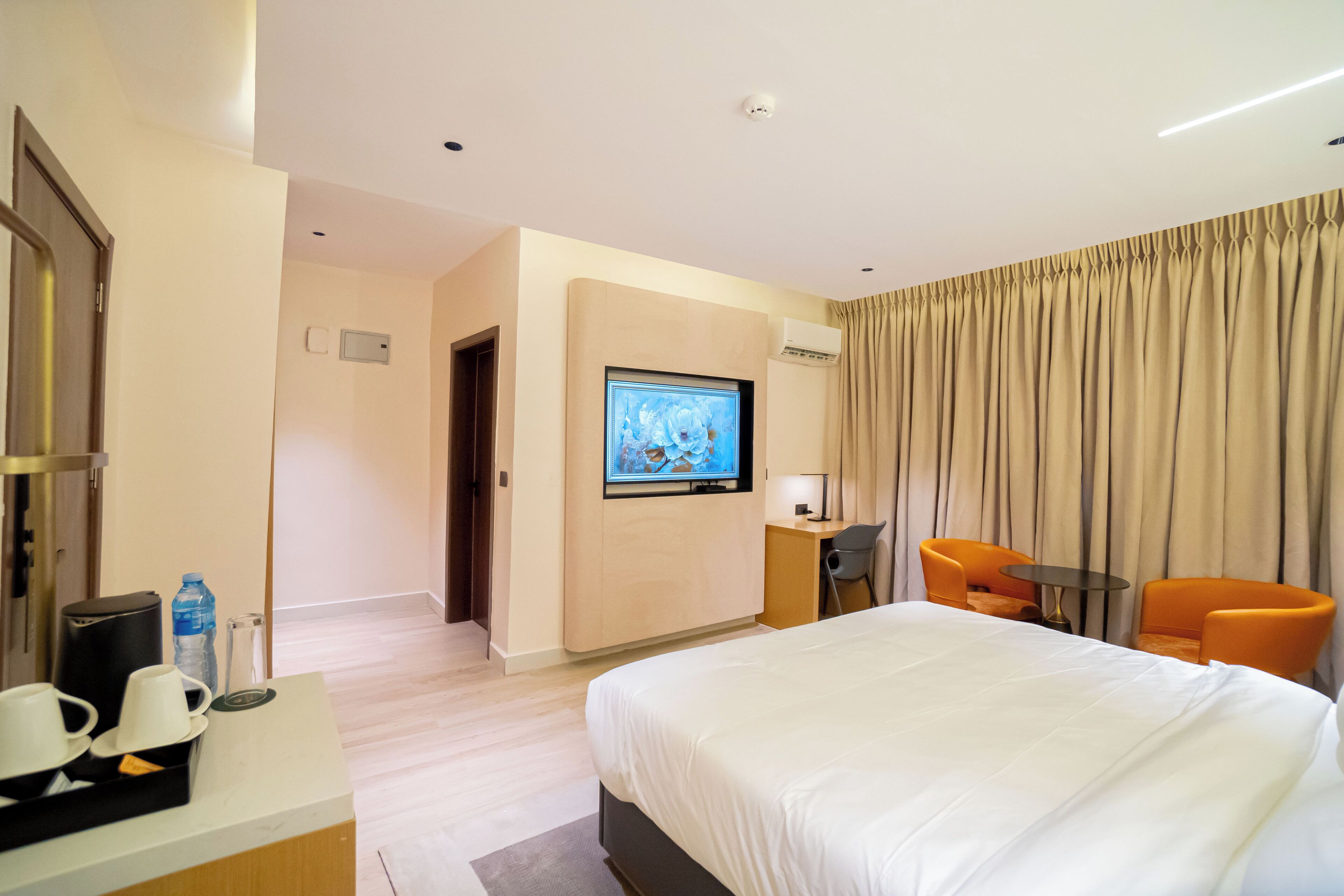 Une chambre d’hôtel avec un grand lit, un téléviseur fixé au mur, un bureau avec une chaise et deux fauteuils orange.