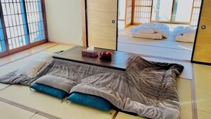 Una habitación con tatami, un futón, una mesita con una caja de pañuelos y un objeto rojo, y un puertas correderas que dan a una habitación tradicional.