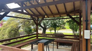 A covered outdoor seating area with benches and a table, surrounded by trees and a view of a park.