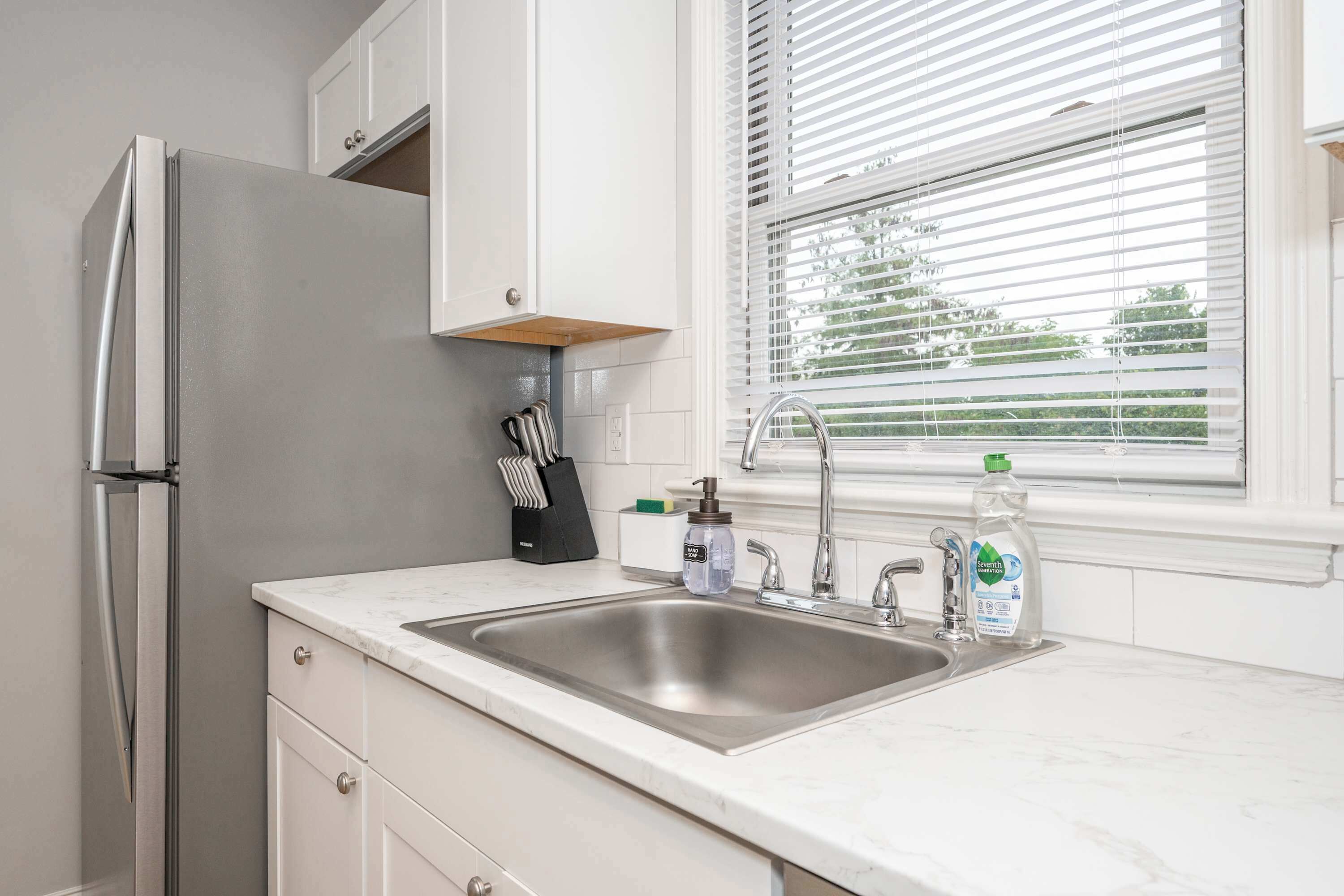 A kitchen with a stainless steel refrigerator, a sink with a faucet, and a window with blinds.