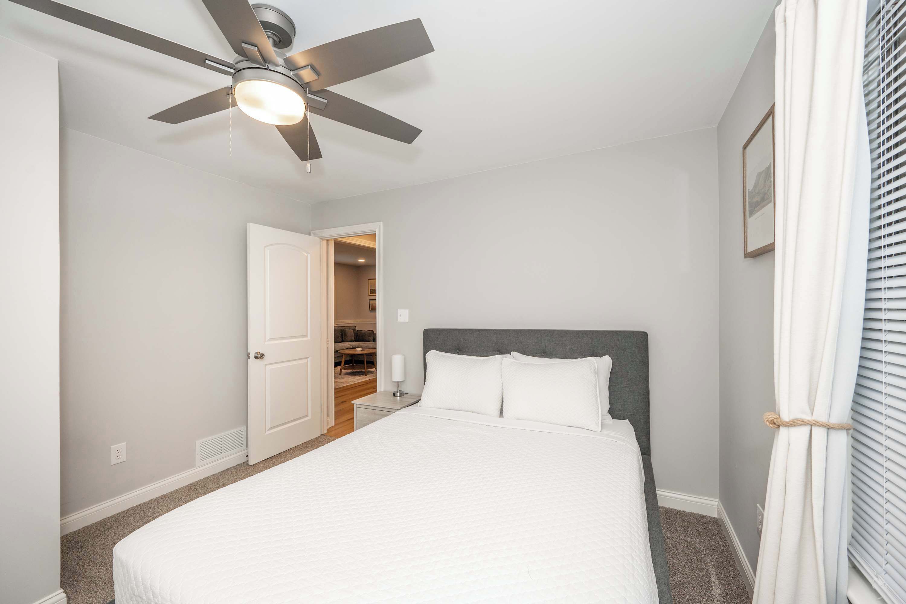 A bedroom with a bed, a ceiling fan, a door leading to a hallway, and a window with blinds.