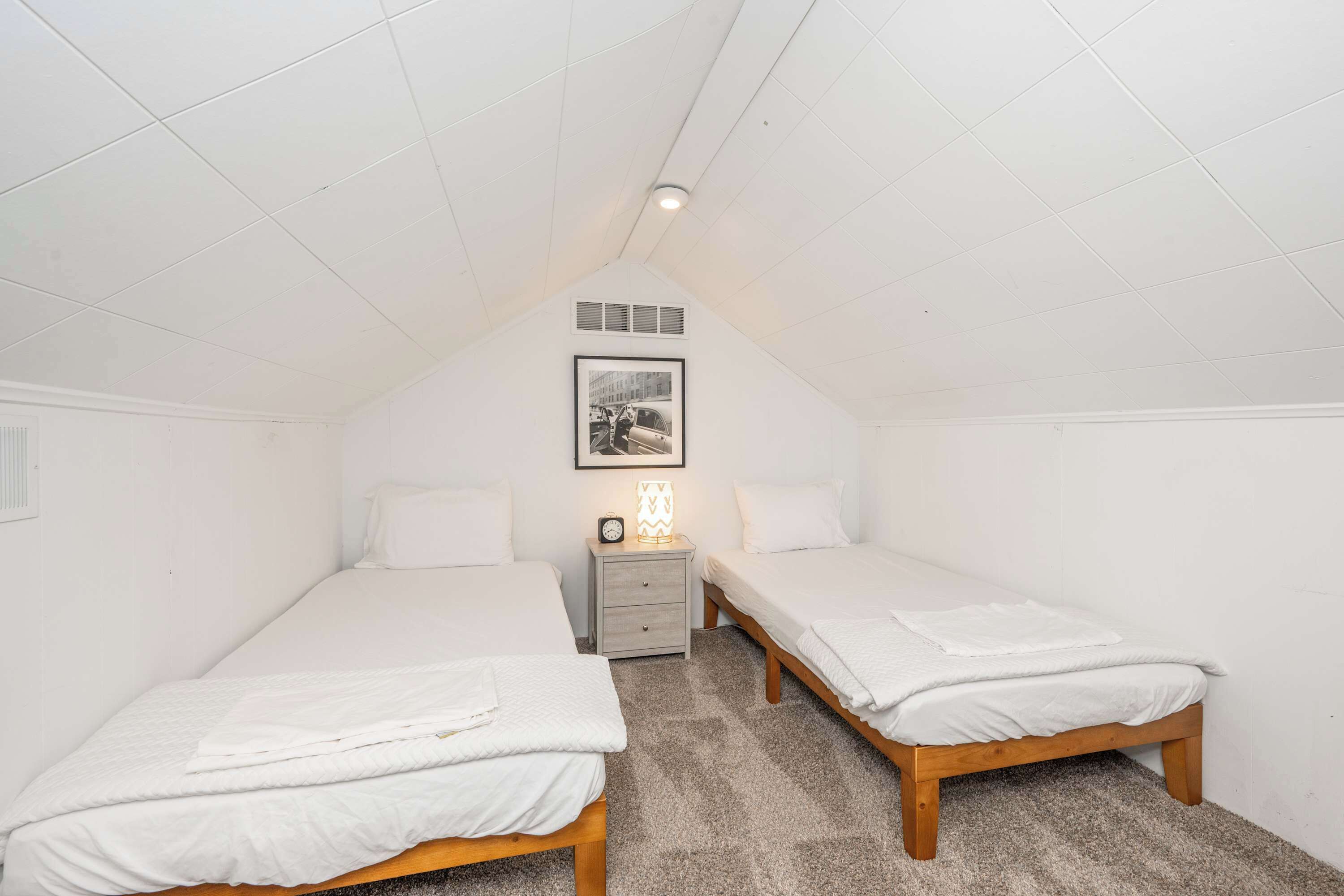 Une petite chambre à coucher de style mansardé, aux murs blancs, avec deux lits simples, une table de chevet avec une lampe et un tableau encadré au mur.