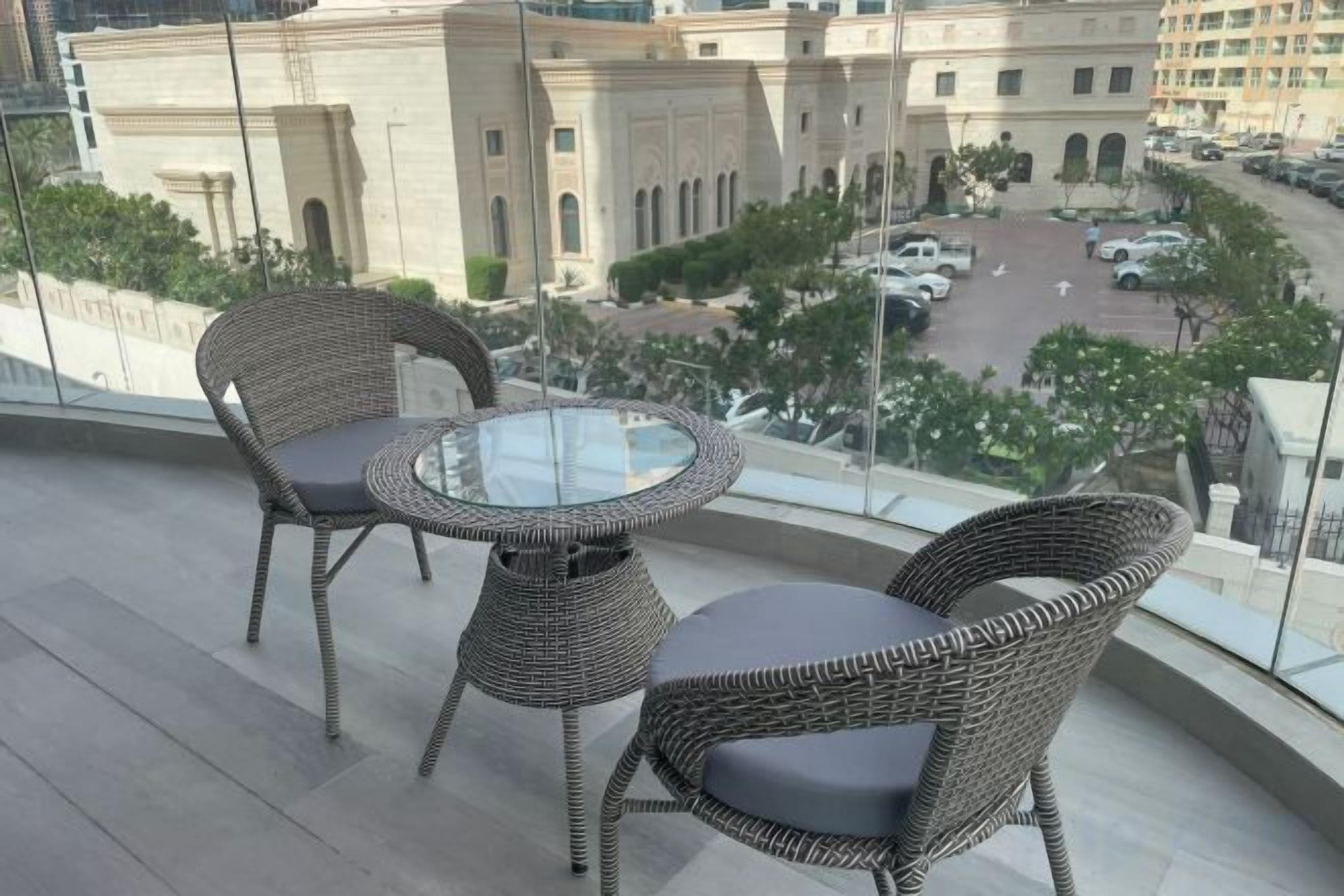 A balcony with wicker furniture and a glass table, overlooking a cityscape with buildings and a parking area.