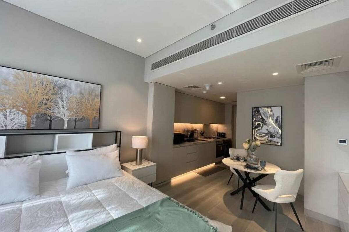 A modern hotel room with a bed, bedside table, lamp, and a dining area with a table and chair.