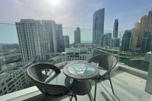A balcony with a glass table and wicker chairs, overlooking a cityscape with modern buildings and a river. - Dubai Marina Walk Gem Modern Apt with Pool & Gym access, Steps to Beach & Metro (Dubai)