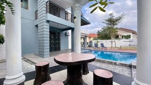 A poolside area with a marble table and stools, surrounded by white columns, and a view of a swimming pool and buildings in the background. - Stylish Blue Water Home w/ Pool (Tebrau)