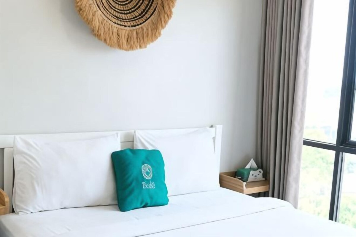 A bed with white linens and a teal pillow, a woven wall hanging above, and a window with curtains to the right.