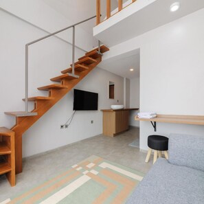A modern interior with a wooden staircase, a flat-screen TV mounted on the wall, a wooden cabinet, and a gray sofa. - Bale Puri (West Jakarta)