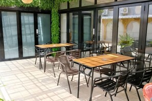 An outdoor seating area with tables and chairs, a tiled floor, and a glass wall with a view. - BALE Puri (West Jakarta)