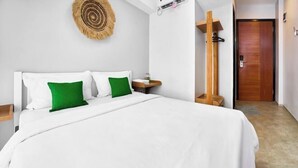 A neatly made bed with white and green pillows, a wooden headboard, a small bedside table with a bottle, and a wooden door in the background. - BALE Puri (West Jakarta)