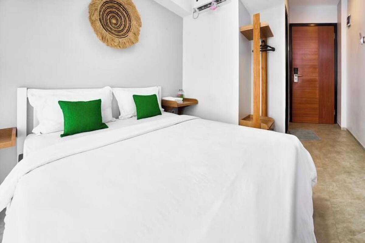 Una cama bien hecha con almohadas blancas y verdes, un cabecero de madera, una mesita de noche con una botella y una puerta de madera al fondo.