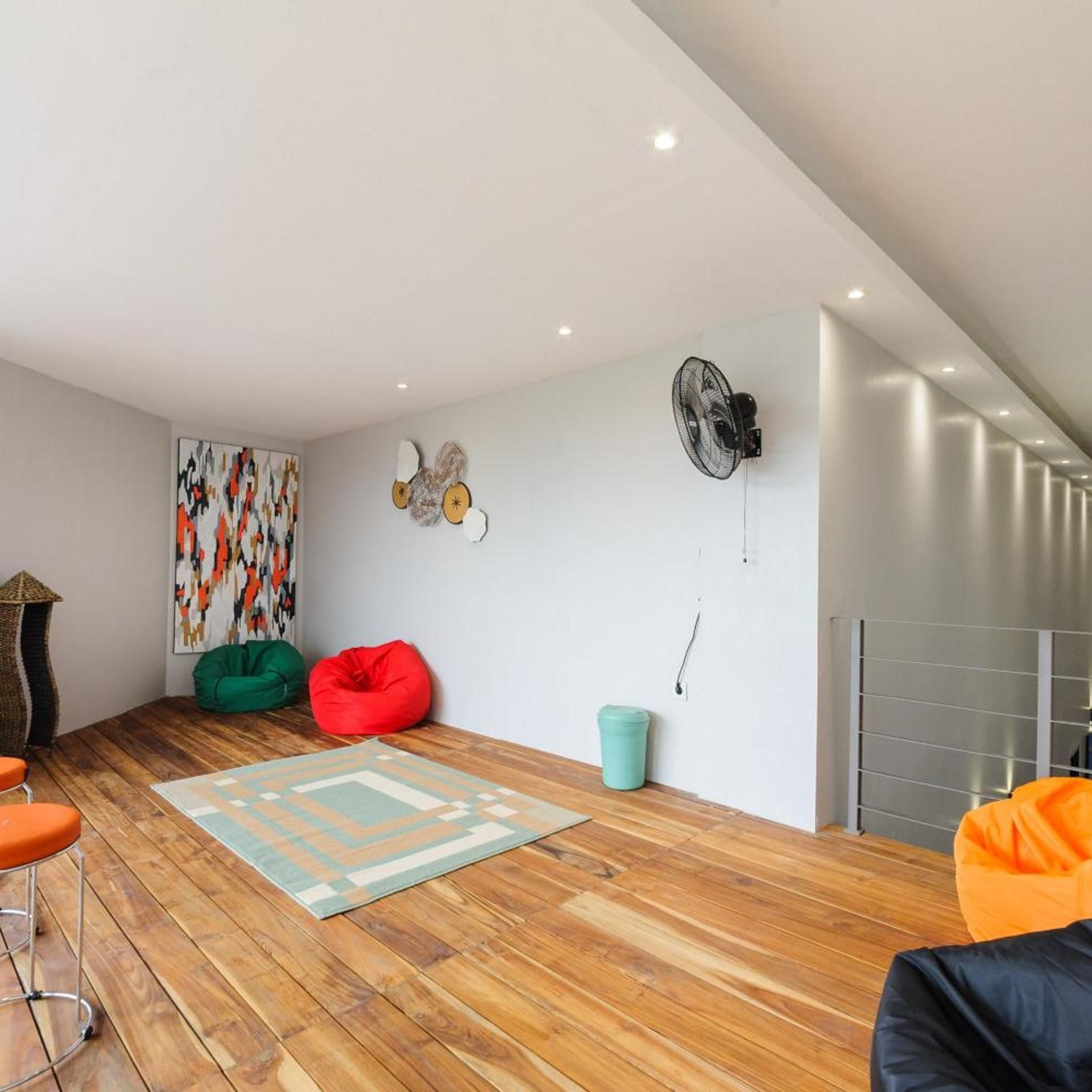 A modern living room with a wooden floor, a colorful abstract painting, a green and red bean bag, a small trash bin, and a fan on the wall.