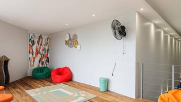 A modern living room with a wooden floor, a colorful abstract painting, a green and red bean bag, a small trash bin, and a fan on the wall.