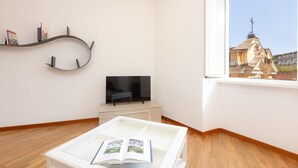 A modern living room with a flat-screen TV, a white coffee table with magazines, a curved wall shelf with books, and a large window with a view of a building. - Rental in Rome Ripa Apartment (Rome)