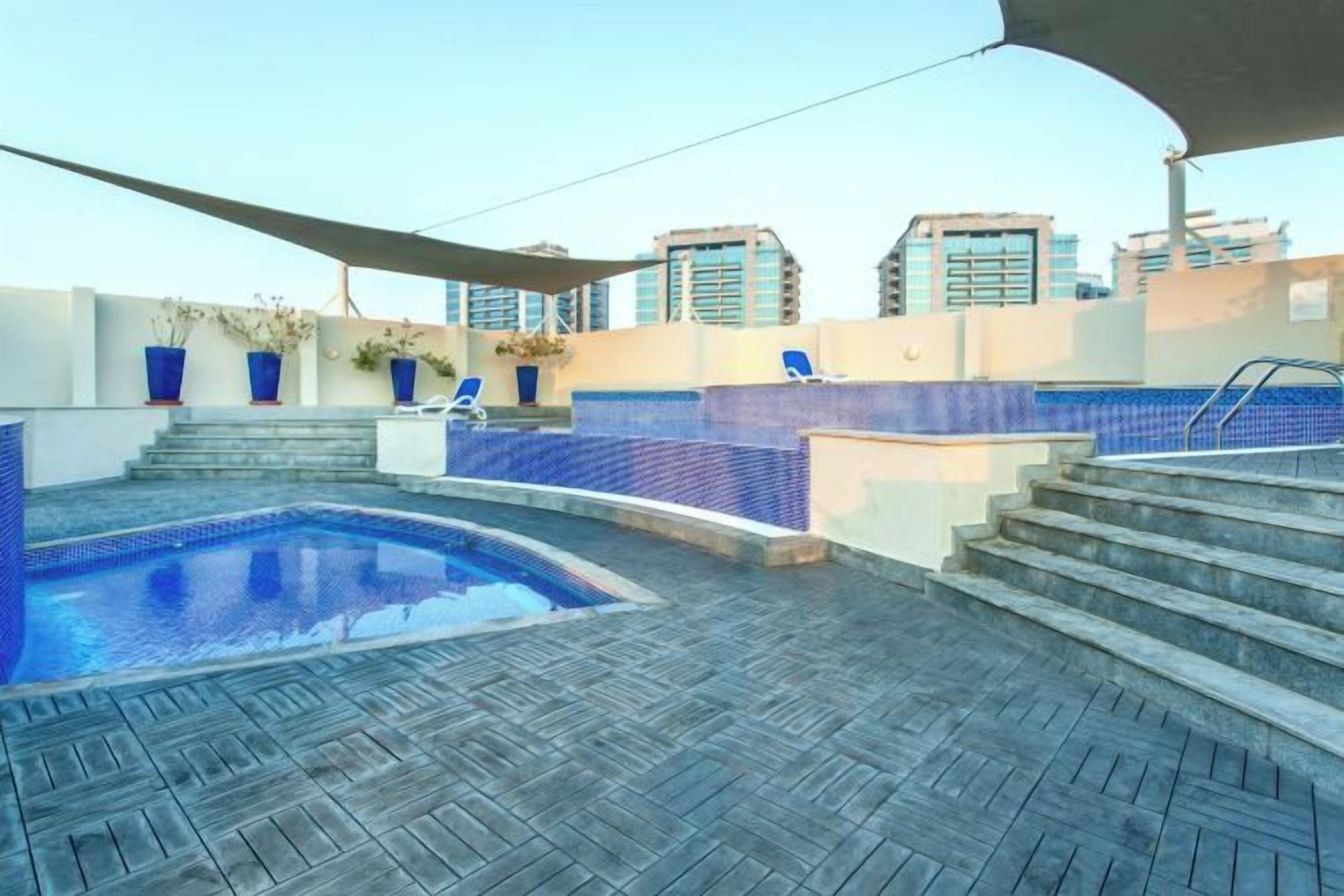 A rooftop swimming pool with steps leading to a lounge area, surrounded by buildings.