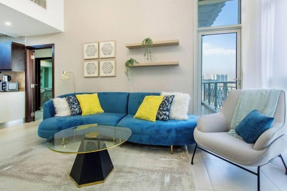 Ein modernes Wohnzimmer mit einem blauen Sofa, einem Glastisch, einem weißen Sessel und einem Balkon mit Aussicht.
