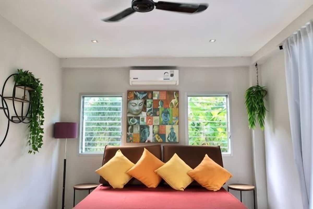 Quarto com cama, ventoinha no teto, aparelho de ar condicionado, duas janelas com cortinas e planta na parede.